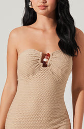 A woman is wearing a strapless, beige textured dress that features a gathered bust with a small opening in the center, decorated with large pearl and marble beads. She has long, dark, wavy hair and is accessorized with large, shiny hoop earrings.